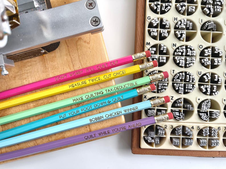 May include: Six hexagonal pencils in pink, yellow, light green, turquoise, light blue, and purple, each with a gold ferrule and pink eraser. The pencils feature the text: DON'T WORRY IT'LL QUILT OUT, MEASURE TWICE, CUT ONCE!, MAKE QUILTING TAX-DEDUCTIBLE, PUT YOUR FOOT DOWN & QUILT, BOBBIN CHICKEN WINNER, and QUILT WHILE YOU'RE AHEAD.
