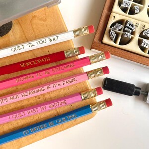 May include: A set of six hexagonal pencils in white, red, dark pink, light pink, magenta, and blue, each featuring gold-stamped text including QUILT 'TIL YOU WILT, SEWCIOPATH, ITCHING TO BE STITCHING, IT'S NOT HOARDING IF IT'S FABRIC, I QUILT PAST MY BEDTIME, and I'D RATHER BE QUILTING.