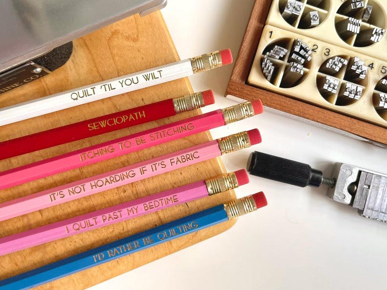 May include: A set of six hexagonal pencils in white, red, dark pink, light pink, magenta, and blue, each featuring gold-stamped text including QUILT 'TIL YOU WILT, SEWCIOPATH, ITCHING TO BE STITCHING, IT'S NOT HOARDING IF IT'S FABRIC, I QUILT PAST MY BEDTIME, and I'D RATHER BE QUILTING.