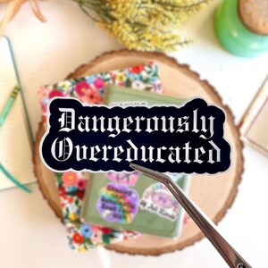 May include: A black sticker with the words "Dangerously Overeducated" in white gothic lettering. The sticker is held by tweezers and rests on a floral fabric and a green box. Other items include a notebook, pencil, and a book.