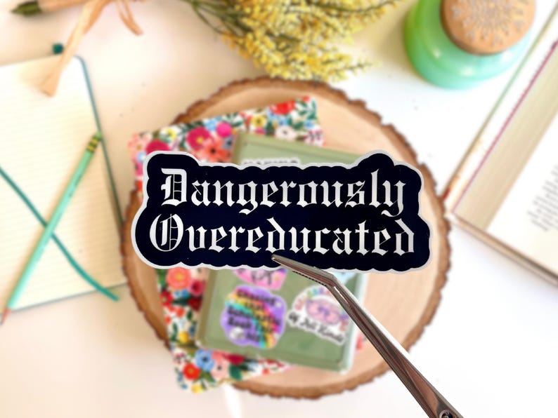 May include: A black sticker with the words "Dangerously Overeducated" in white gothic lettering. The sticker is held by tweezers and rests on a floral fabric and a green box. Other items include a notebook, pencil, and a book.