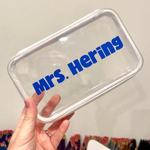 May include: A clear, rectangular cosmetic bag with a white trim. The words "MRS. Hering" are printed in bold blue letters across the front of the bag. The bag is held up against a light background.