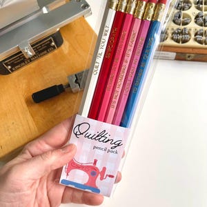 May include: A pack of six pencils in a clear plastic sleeve with a label reading Quilting pencil pack and a sewing machine graphic. The pencils are white, red, dark pink, light pink, magenta, and blue, each with gold text and a metal ferrule.