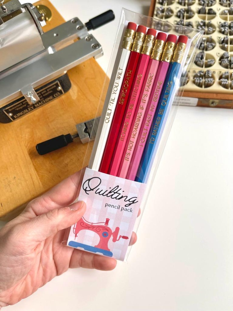May include: A pack of six pencils in a clear plastic sleeve with a label reading Quilting pencil pack and a sewing machine graphic. The pencils are white, red, dark pink, light pink, magenta, and blue, each with gold text and a metal ferrule.