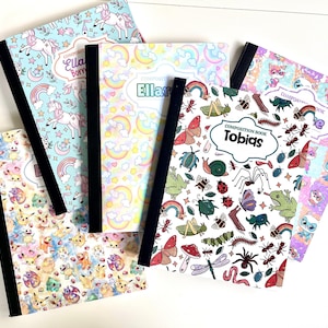 May include: Four composition books with black spines and various colorful covers. One features unicorns, one has rainbows, one displays insects and the name Tobias, and one shows cartoon characters. Each book has a white label for personalization.