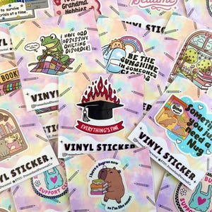 May include: A collection of colorful vinyl stickers with various designs, including a frog, a graduation cap, a cat, and a bag. The stickers have text like "Everything's Fine" and "Sometimes you just need a Nug".