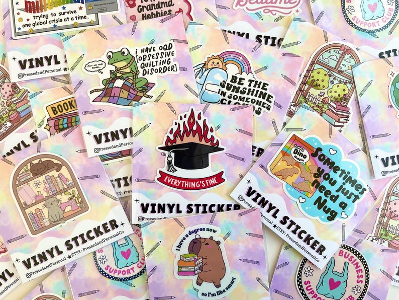 May include: A collection of colorful vinyl stickers with various designs, including a frog, a graduation cap, a cat, and a bag. The stickers have text like "Everything's Fine" and "Sometimes you just need a Nug".