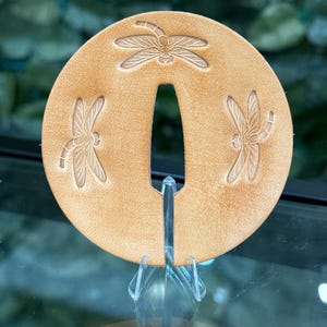 May include: A tan leather decorative piece with three dragonfly designs. The piece has a central opening and is displayed on a clear stand. The dragonflies are embossed on the leather.