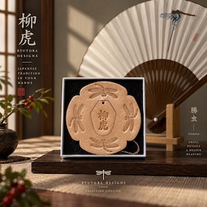 May include: A tan, circular, embossed leather-like ornament featuring four dragonfly motifs surrounding a central hexagonal plaque with the characters 柳虎. The item is presented in a square white box with a black interior lining.