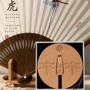 Puede incluir: Un objeto circular de cuero color canela con motivos de libélulas en relieve y una etiqueta rectangular central con los caracteres 柳虎, dentro de una caja blanca. Detrás hay un abanico plegable con una ilustración de libélula y el texto RYUTORA DESIGNS.