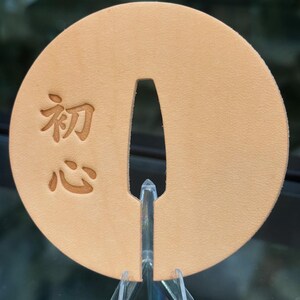 May include: A round, tan leather object with a central slot and Japanese characters. The object is displayed on a clear stand. The Japanese characters are "初心", which translates to "beginner's mind".