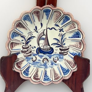 May include: A scalloped ceramic plate featuring a hand-painted sailboat in the center, surrounded by blue and brown floral motifs. The rim is decorated with alternating blue and brown grid patterns, and the edge is finished with a light pink trim.