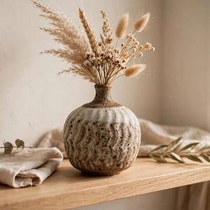 May include: A round, textured ceramic vase with a speckled beige and brown finish, featuring vertical ribbed patterns. The vase holds a dried floral arrangement consisting of pampas grass, wheat stalks, and small fluffy bunny tail grasses.
