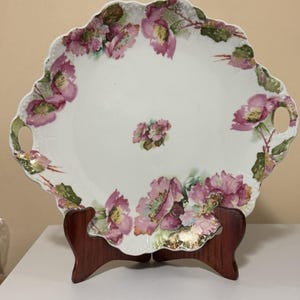 May include: A white porcelain serving platter with a scalloped edge and two integrated handles. The platter features a floral pattern of pink flowers with green leaves and gold accents around the rim and a small floral cluster in the center.