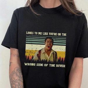 以下が含まれることがあります： ヴィンテージスタイルのグラフィックが特徴の黒いTシャツ。グラフィックには、口を開けた男性の画像と「LOOKS TO ME LIKE YOU'RE ON THE WRONG SIDE OF THE RIVER」というテキストが含まれています。