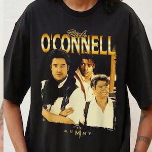 以下が含まれることがあります： 3人の男性のグラフィックと、その上に金色の文字で「Rick O'Connell」と書かれた黒いTシャツ。「The Mummy」の文字は、男性の下に白い文字で書かれています。