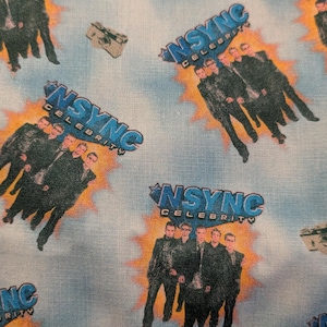 May include: Light blue fabric with a repeating pattern featuring the band *NSYNC*. The design includes images of the band members, the words "NSYNC Celebrity", and vintage cameras. The background has a cloudy sky effect.