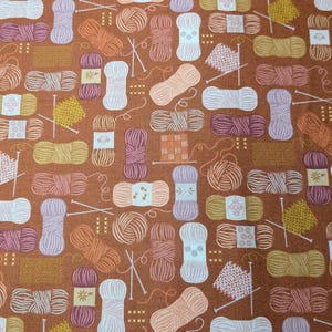 May include: A brown fabric featuring a repeating pattern of yarn balls in shades of pink, purple, yellow, and white, interspersed with small illustrations of knitting needles and knitted swatches, creating a dense and organized textile design.