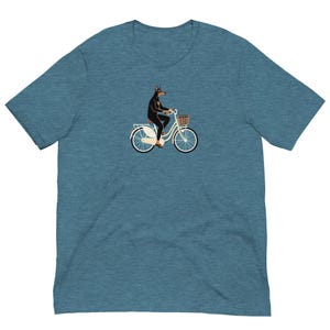 Könnte beinhalten: Türkisfarbenes T-Shirt mit einer Illustration eines Dobermanns auf einem weißen Fahrrad mit Korb. Der Hund trägt ein schwarzes Outfit. Das Fahrrad hat cremefarbene Reifen und einen braunen Korb. Das T-Shirt hat kurze Ärmel.