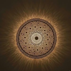 Può includere: Una lampada da parete circolare con un intricato motivo a mandala tagliato al laser in marrone scuro. La fonte di luce dietro il design traforato emette una luce calda e crea un effetto d'ombra radiante sulla parete circostante.
