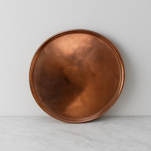 May include: A large, circular copper tray with a perforated surface. The tray has a raised rim and a warm, reddish-brown colour. The tray is likely used for serving or decorative purposes.