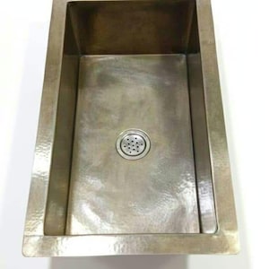 Puede incluir: Un fregadero de cocina rectangular hecho de metal martillado con un acabado cálido en tono bronce parduzco. El fregadero presenta un borde ancho y plano y un conjunto de desagüe circular centrado con un colador de metal perforado sobre fondo blanco.