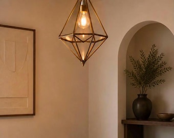 May include: A geometric pendant light fixture with a diamond-shaped metal frame in a brass finish. The fixture hangs from a black cord and ceiling canopy, enclosing a single exposed Edison-style light bulb that emits a warm, soft glow.