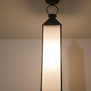 May include: A black, rectangular pendant light fitting with a white, illuminated panel. The light hangs from a circular ceiling mount and features a decorative ring. The fitting is a modern design.