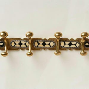 Puede incluir: Un perchero de pared horizontal de latón con cuatro ganchos redondeados. La barra central está decorada con tres incrustaciones rectangulares, cada una con un patrón repetitivo de semicírculos color crema sobre fondo negro, enmarcado por un borde de cuentas.