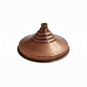 Pode incluir: Um chuveiro de cor cobre com uma textura martelada. O chuveiro tem uma forma circular, larga e rasa, com uma série de anéis concêntricos que conduzem a um encaixe roscado de latão. O design é rústico e decorativo.