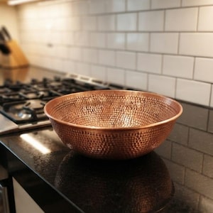 May include: A round, wide-rimmed bowl crafted from polished copper with a distinct hammered texture across its entire exterior and interior surface. The bowl features a warm, metallic reddish-brown finish and a smooth, rounded edge along the top rim.