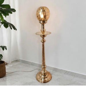 May include: A tall, gold-coloured floor lamp featuring a spherical, perforated metal shade that emits a warm glow. The lamp has a slender, twisted metal stem, a circular tray-like middle section, and a wide, stable circular base made of polished metal.