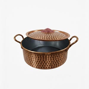 Op de afbeelding: Koperen kookpot met een deksel. De pot heeft een gehamerde textuur en twee handvatten. Het deksel is gedeeltelijk open en onthult het donkere interieur. Het deksel heeft een decoratief rood accent.