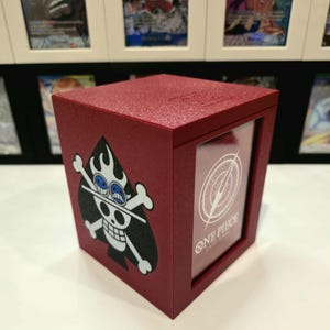 Könnte beinhalten: Burgunderfarbene Kartenaufbewahrungsbox mit einer schwarz-weißen Totenkopf- und gekreuzten Knochen-Grafik und einem blauäugigen Totenkopf. Die Box hat ein klares Fenster, das eine Karte mit dem Text "ONE PIECE CARD GAME" anzeigt.