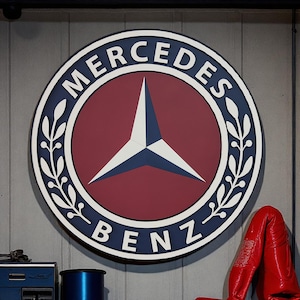 May include: A large, circular Mercedes-Benz logo sign. The sign features a red center with a white and blue star emblem. The words "MERCEDES" and "BENZ" are in white lettering around the edge, with a blue border and white leaf accents.