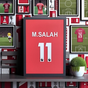 May include: Framed red artwork featuring the name "M. SALAH" and the number "11" in white. The artwork is surrounded by framed images of soccer jerseys and trophies. The Premier League logo is at the bottom of the number 11.