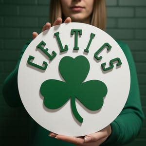 May include: A circular white sign featuring the word "CELTICS" in green lettering and a green shamrock. The sign is held up in front of a green brick wall.