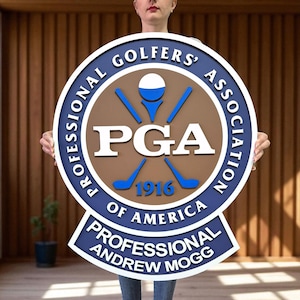 Può includere: Un cartello rotondo blu, marrone e bianco con la scritta "PROFESSIONAL GOLFERS' ASSOCIATION OF AMERICA" e "PGA 1916". Il cartello presenta una pallina e mazze da golf. Il cartello è tenuto da una persona.