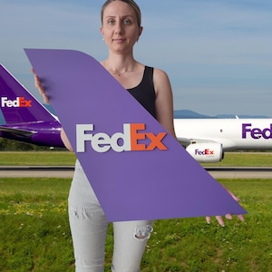 May include: A purple FedEx sign with white and orange lettering is held in front of a FedEx airplane. The sign is in the shape of an airplane wing. The background includes a grassy field and a blue sky.