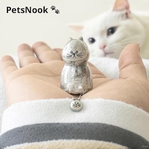 Peut inclure: Une petite figurine de chat grise avec des détails faciaux noirs est tenue dans la main d'une personne. La figurine a un pendentif assorti. Le texte "PetsNook" est en haut à gauche de l'image, avec des empreintes de pattes à côté. Un chat blanc est en arrière-plan.