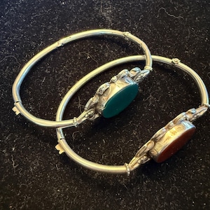 May include: Two silver-toned hinged bangle bracelets, each featuring a central oval-shaped gemstone. One bracelet holds a smooth, dark green stone, while the other contains a smooth, reddish-brown stone. Both bangles have decorative metalwork.