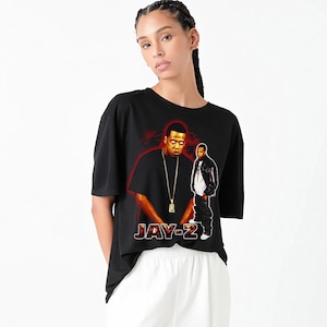May include: Black t-shirt with a graphic of Jay-Z in two poses and the text "JAY-Z". The shirt has a crew neck and short sleeves. The graphic has red and orange accents.