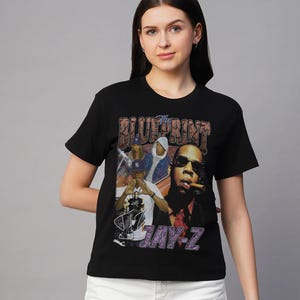 May include: Black t-shirt with a retro graphic of Jay-Z, the text "The Blueprint" and "JAY-Z". The design includes images of Jay-Z and other figures. The shirt has a crew neck and short sleeves.