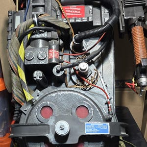 May include: A detailed, black Ghostbusters proton pack prop with various tubes, wires, and canisters. The prop features a circular component with red accents and a warning label. The dimensions are approximately 24.5 inches high, 14.25 inches wide, and 18 inches deep.
