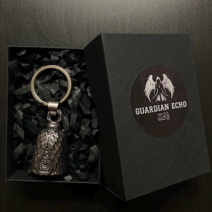 Op de afbeelding: Een zilverkleurige bel met een uilontwerp, bevestigd aan een sleutelhanger, gepresenteerd in een open zwarte doos met versnipperd zwart papier. De doos heeft een rond label met de tekst "GUARDIAN ECHO" en een gevleugeld embleem.