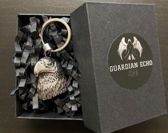 May include: A silver-colored eagle head keychain inside a black gift box with black crinkle paper. The box has a round sticker with the words "GUARDIAN ECHO" and a winged design. The keychain is made of metal.