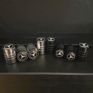 May include: Eight black and silver valve stem caps are displayed. Each cap has a silver band and a white emblem with a three-pointed star. The cylindrical caps are likely made of metal and are arranged in a row.