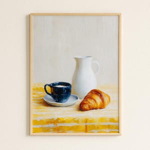 Op de afbeelding: Een ingelijst stilleven schilderij met een donkerblauwe kop en schotel, een witte kan en een gouden croissant op een gele tafelkleed. Het schilderij heeft een lichtbeige achtergrond en een houten lijst.