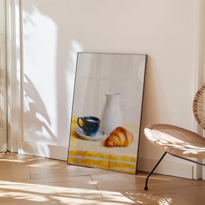 May include: A framed still life painting depicts a breakfast scene. A blue cup and saucer, a white pitcher, and a golden croissant rest on a yellow and white checkered tablecloth. The artwork is leaning against a white wall.