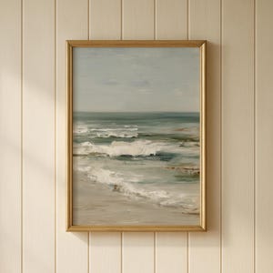 May include: A framed oil painting depicts a seascape with a muted color palette. The artwork features a sandy beach, foamy waves, and a calm ocean under a cloudy sky. The frame is a warm gold color.